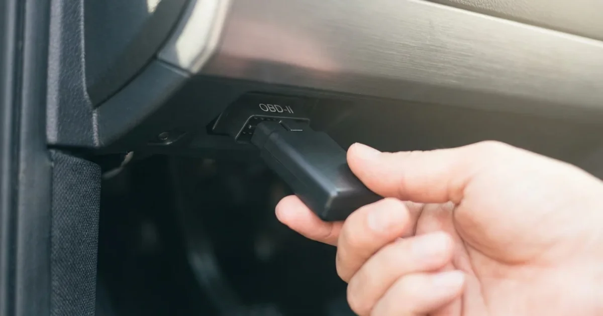 Close-up of a hand connecting a black OBD-II device into a vehicle's diagnostic port.