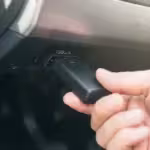 Close-up of a hand connecting a black OBD-II device into a vehicle's diagnostic port.