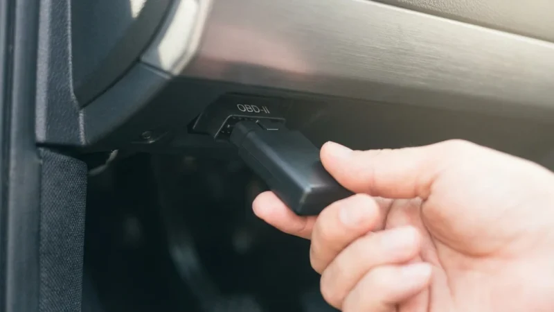 Close-up of a hand connecting a black OBD-II device into a vehicle's diagnostic port.