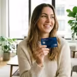 A smiling young adult holds a new bank card in a bright, modern living room.
