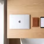 Top-down view of closed silver MacBook, brown leather wallet, and smartphone on wooden desk
