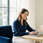 A professional woman smiling confidently while working on her laptop in a bright, modern home office.