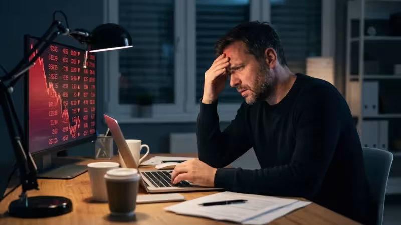 A man looking stressed in a dark room with red light from a financial screen reflecting on his face.