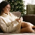Woman in cream sweater dress using smartphone on sofa with Christmas tree and shopping bags.